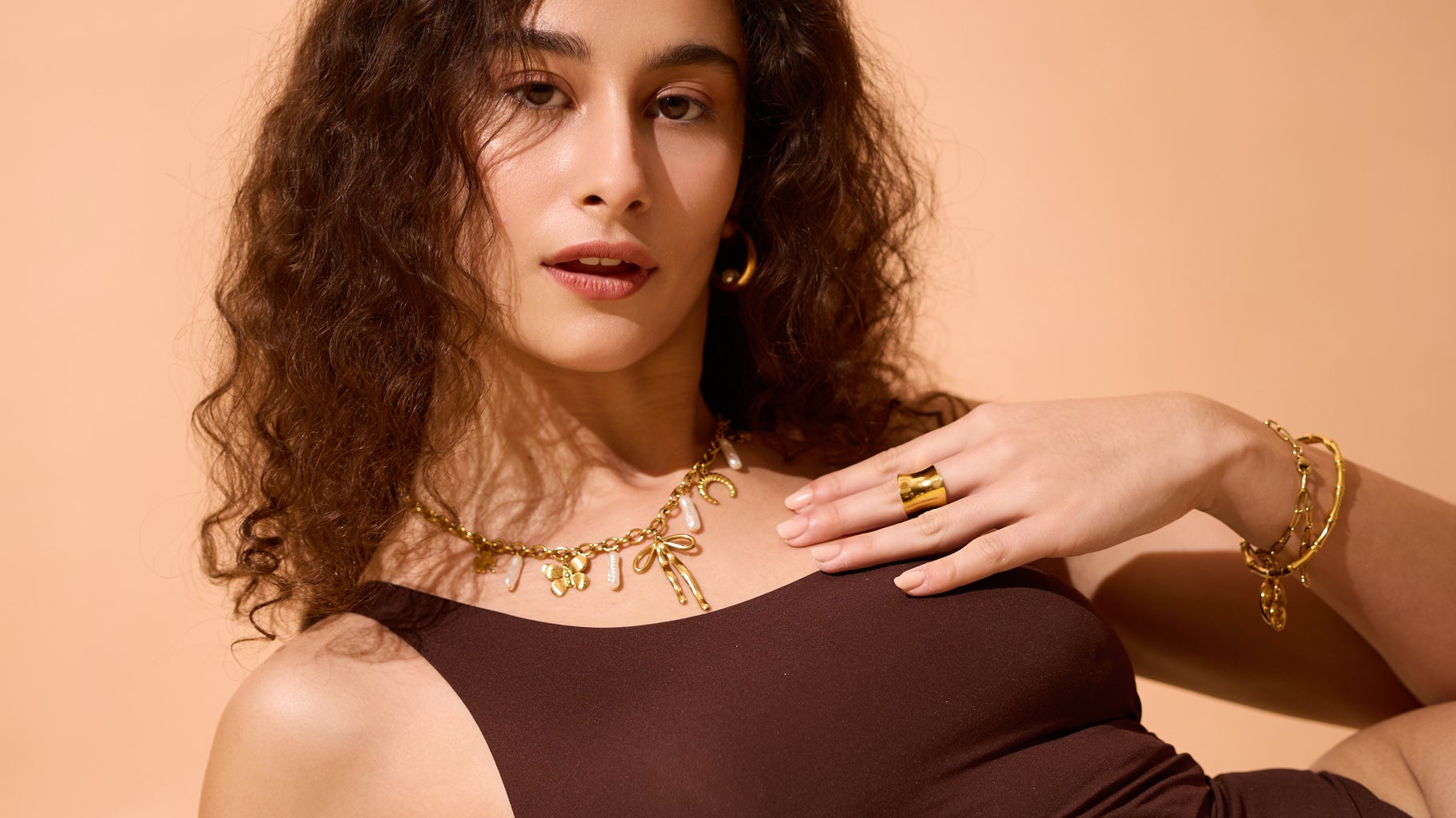 Woman wearing a layered 18K gold-plated charm necklace with pearls, bold gold ring, hoop earrings, and matching bracelets, styled with a brown sleeveless top against a warm beige background.