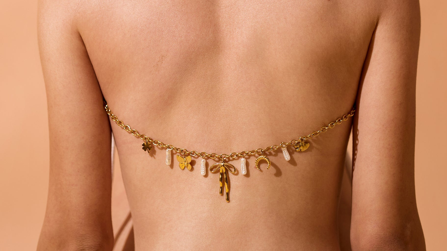 Back view of a woman wearing a delicate 18K gold-plated charm chain with mixed pendants and pearls styled across her back, set against a warm beige background.