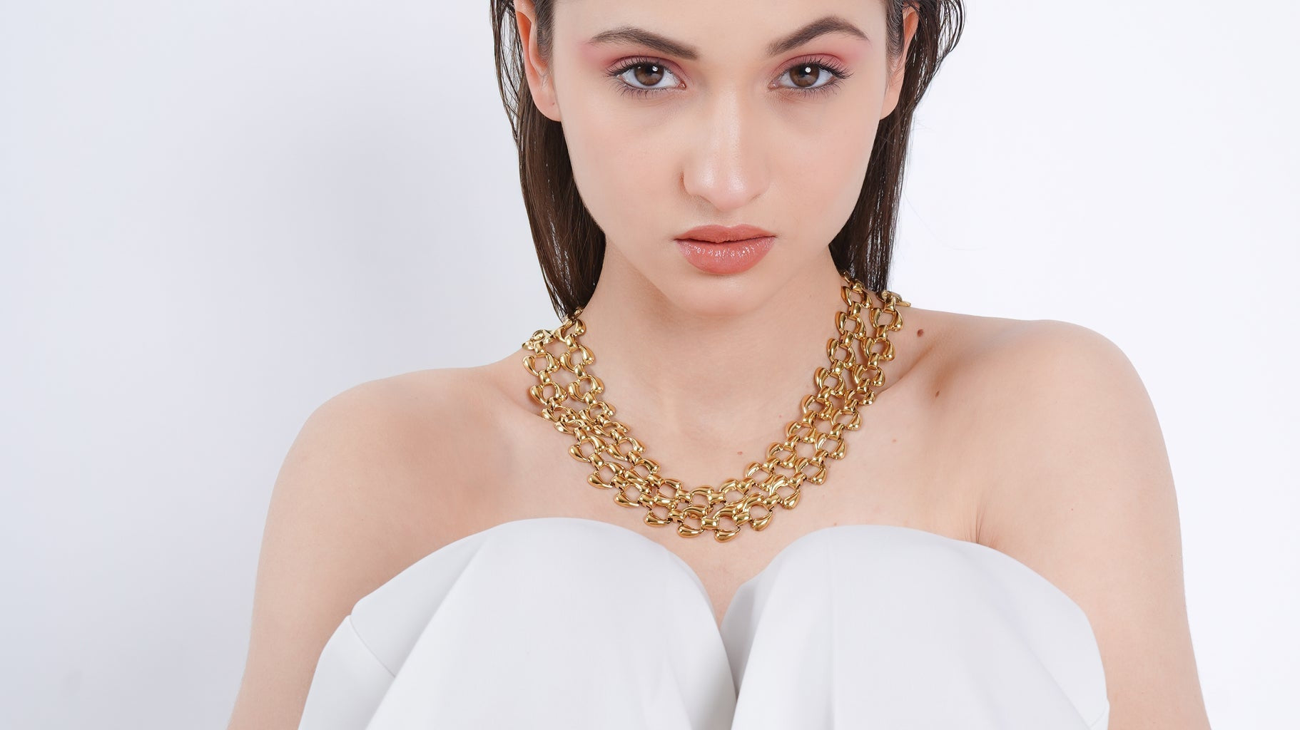 Model wearing a bold 18K gold-plated statement chain necklace with interlinked oval links, styled against a white strapless outfit on a minimal background.