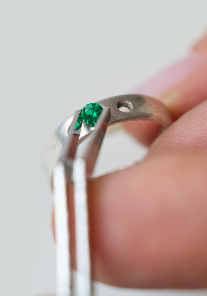 A jeweler carefully setting a vibrant green gemstone into a silver ring using precision tweezers. The close-up captures the delicate craftsmanship and attention to detail in the jewelry-making process.