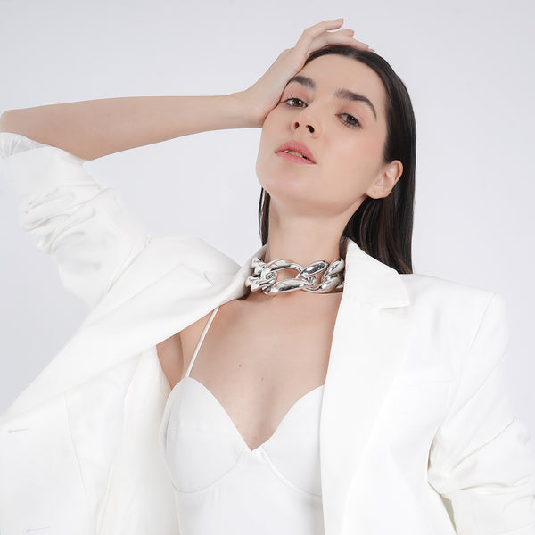 Woman wearing a bold, chunky silver plated chain choker necklace paired with a white structured blazer and a white bustier-style top. Her hair is sleek and straight, and she poses confidently with one hand resting on her forehead against a plain light background.