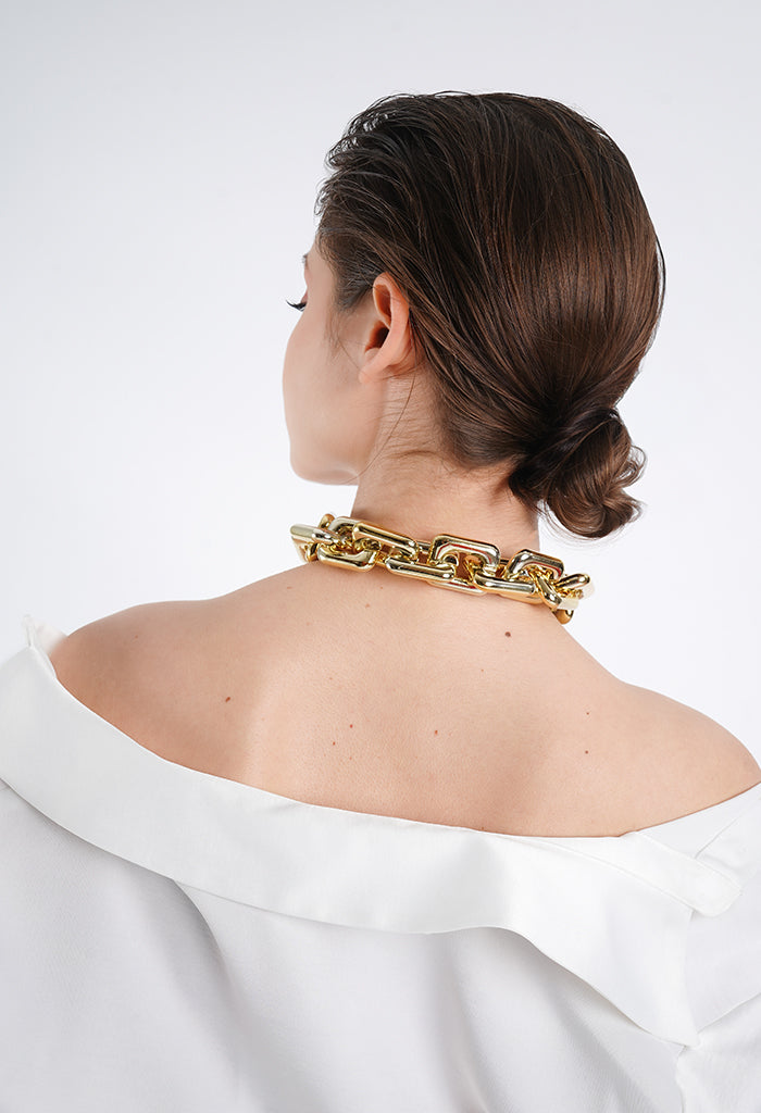 A woman wearing an 18K gold-plated chunky chain necklace with bold, oversized links. The statement piece sits elegantly on her neck, complementing her sleek, pulled-back hairstyle and off-shoulder white outfit, exuding modern sophistication and confidence.