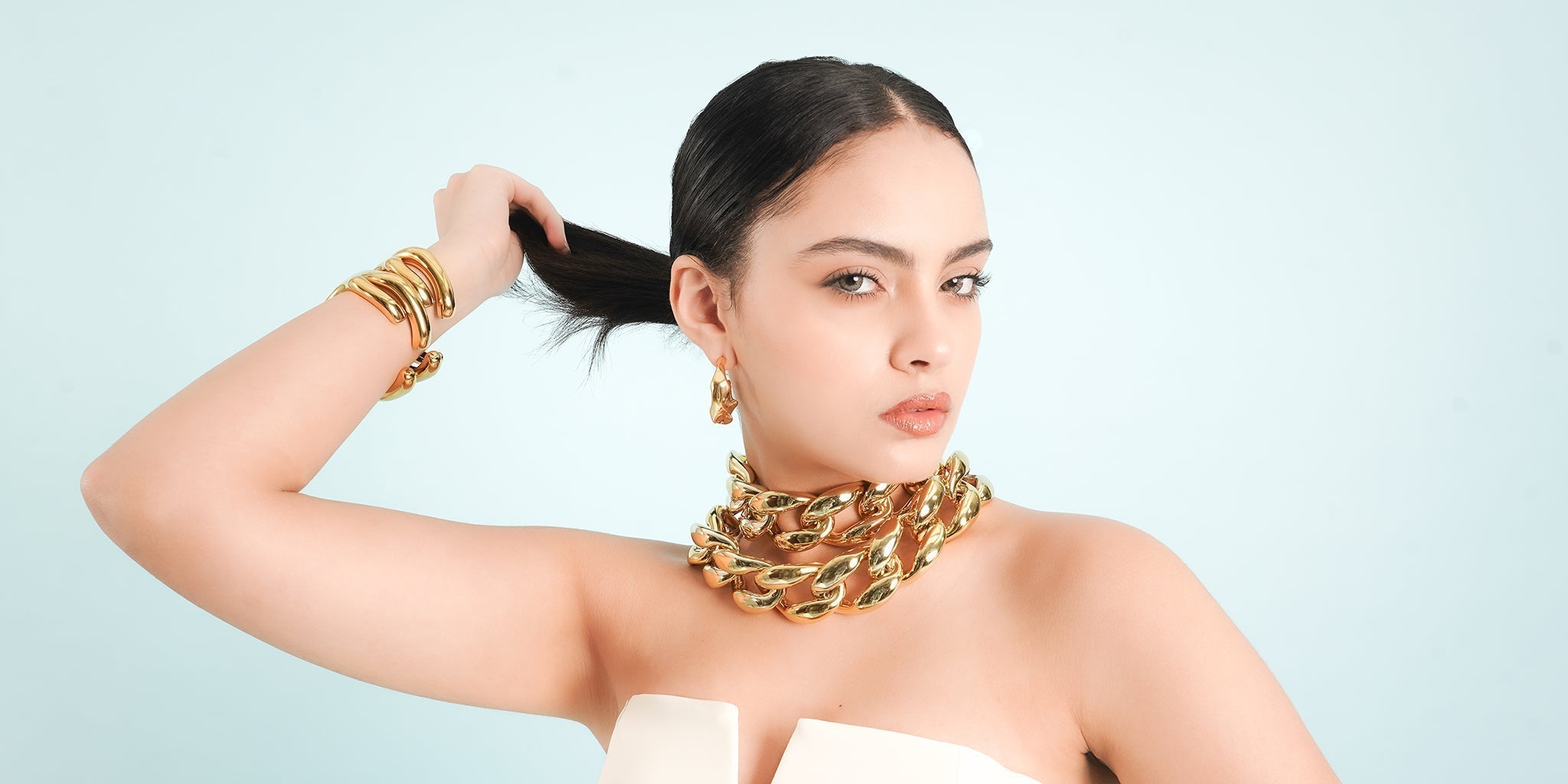 Model wearing bold 18K gold-plated chunky chain necklace, bracelets, and hoop earrings, styled with a sleek ponytail against a soft pastel backdrop—showcasing 2025 jewelry trends by Embrell.
