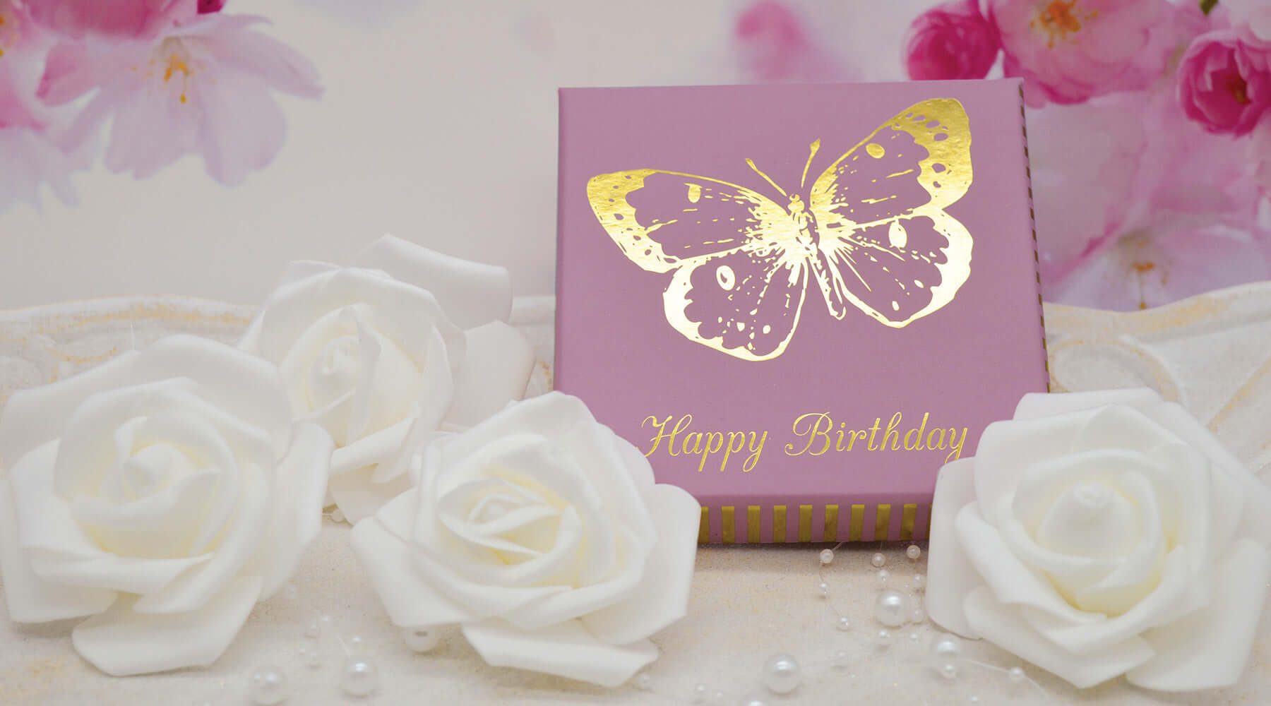 A purple gift box with a gold foil butterfly design and "Happy Birthday" written in elegant script, surrounded by white roses and pearl-like beads. A soft floral background adds a delicate, celebratory touch.