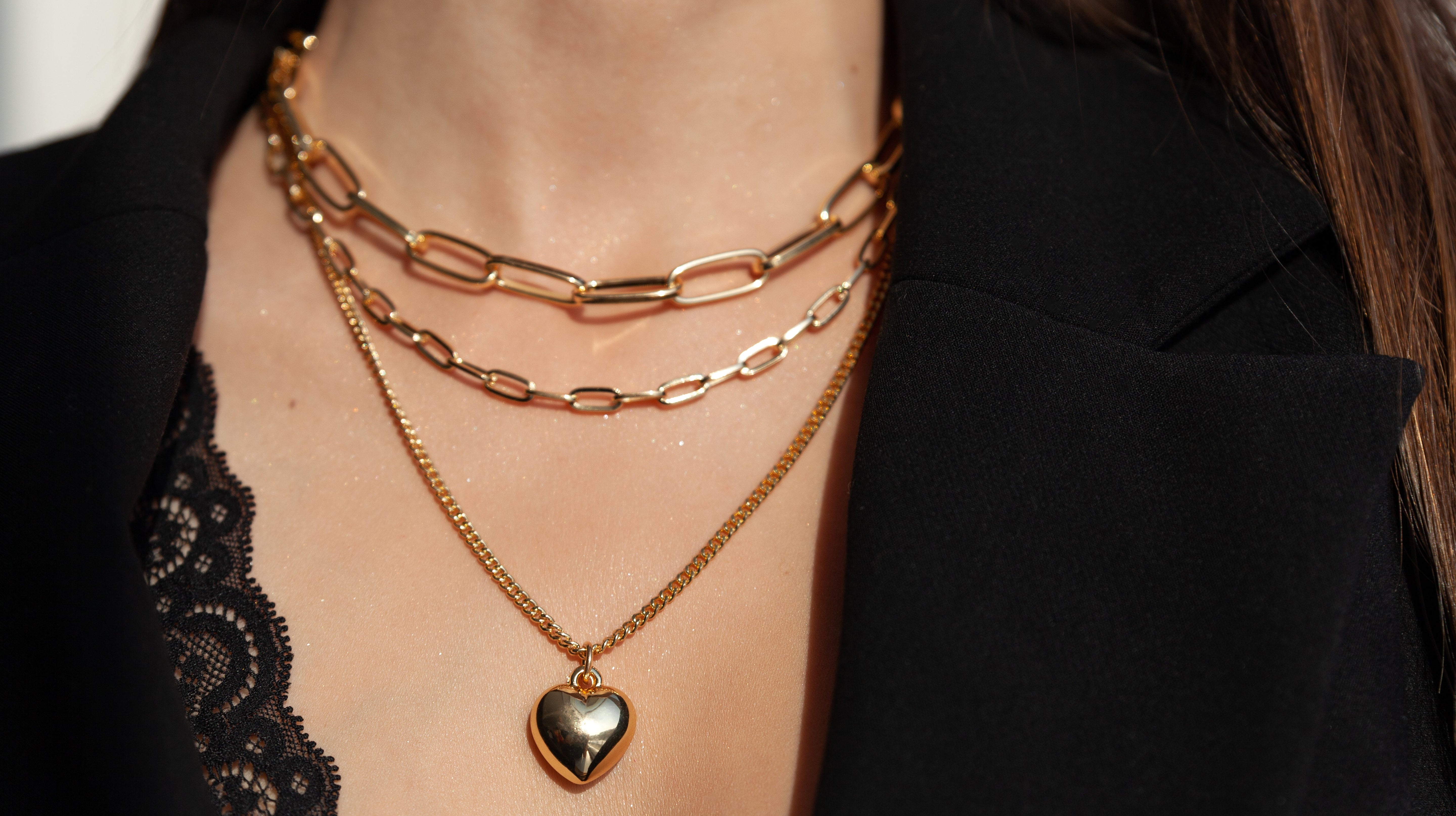 Close-up of layered 18K gold-plated necklaces, including a heart pendant and chain links, styled over a black lace top and blazer.