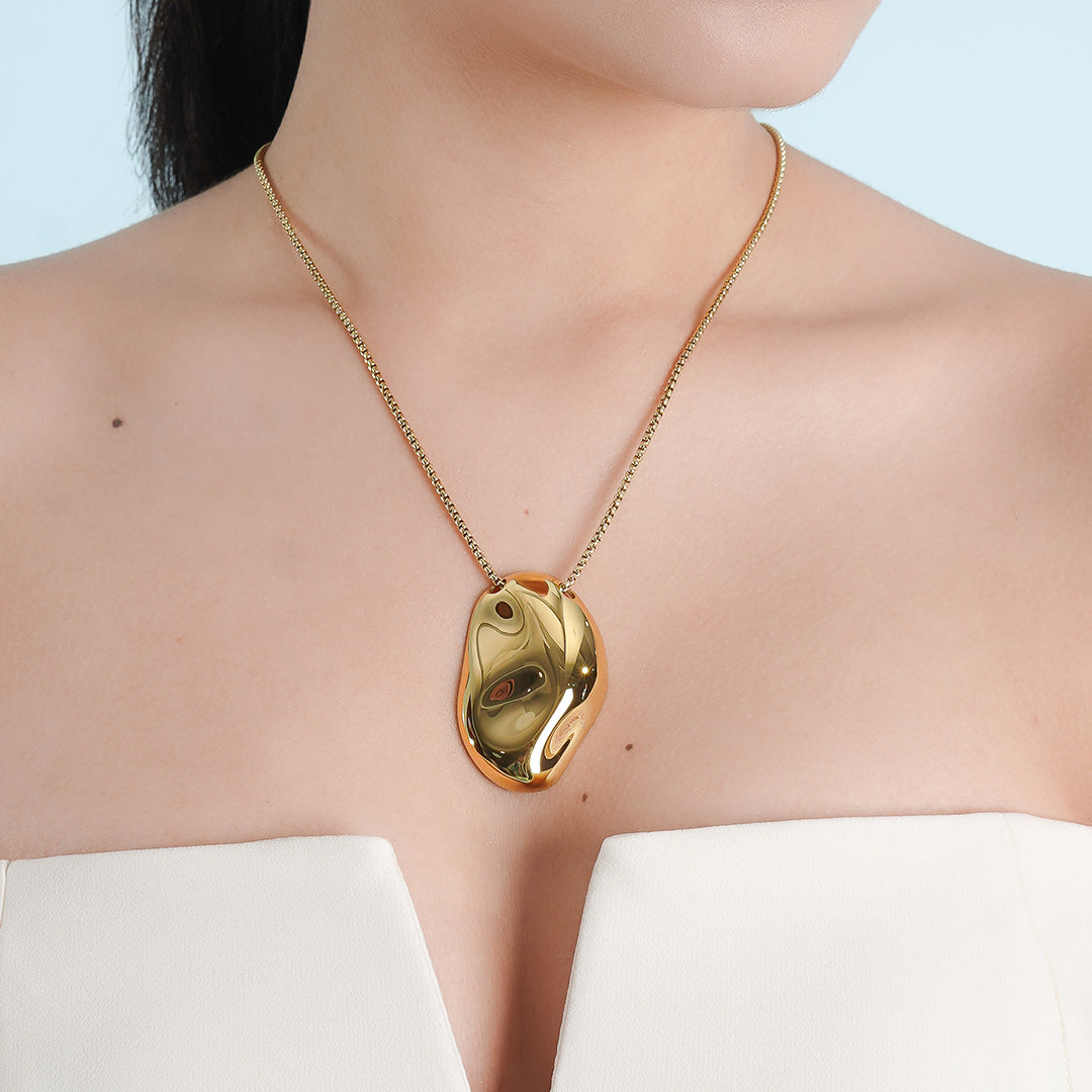 A woman wearing a strapless white outfit showcases a bold 18K gold-plated pendant necklace with an abstract, organic shape. The statement piece reflects light elegantly, enhancing its luxurious appeal.