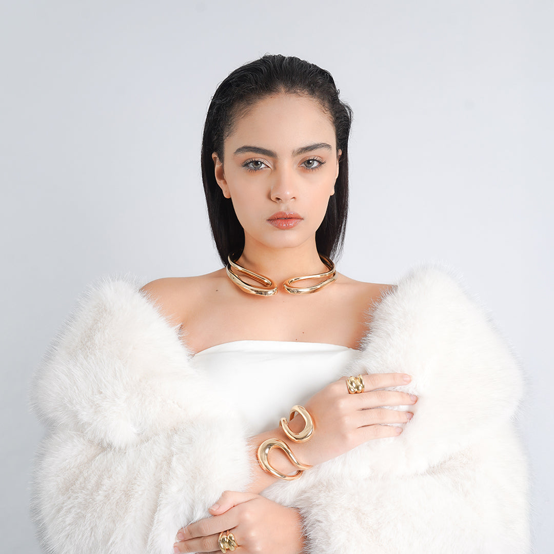 A model elegantly showcasing a luxurious gold jewelry ensemble, featuring a bold statement collar necklace, matching cuff bracelets with fluid curves, and a gold ring with a textured design.