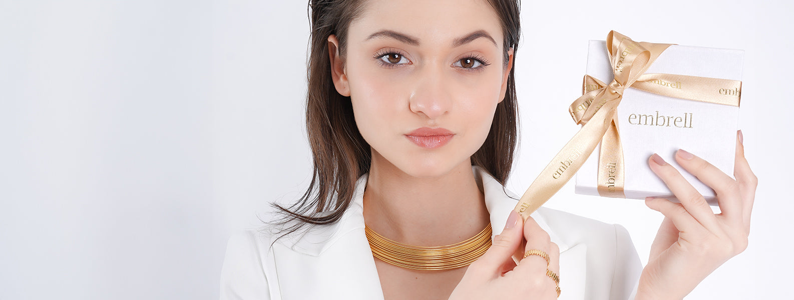 Model wearing an 18K gold-plated layered choker and delicate rings, elegantly holding an Embrell jewelry gift box wrapped with a gold ribbon.