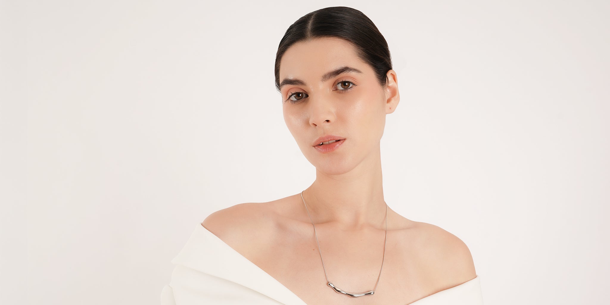 Woman with sleek, center-parted hair wearing an off-shoulder white outfit and a minimalist silver necklace featuring a curved bar pendant. She poses against a plain white background with a soft, natural makeup look.