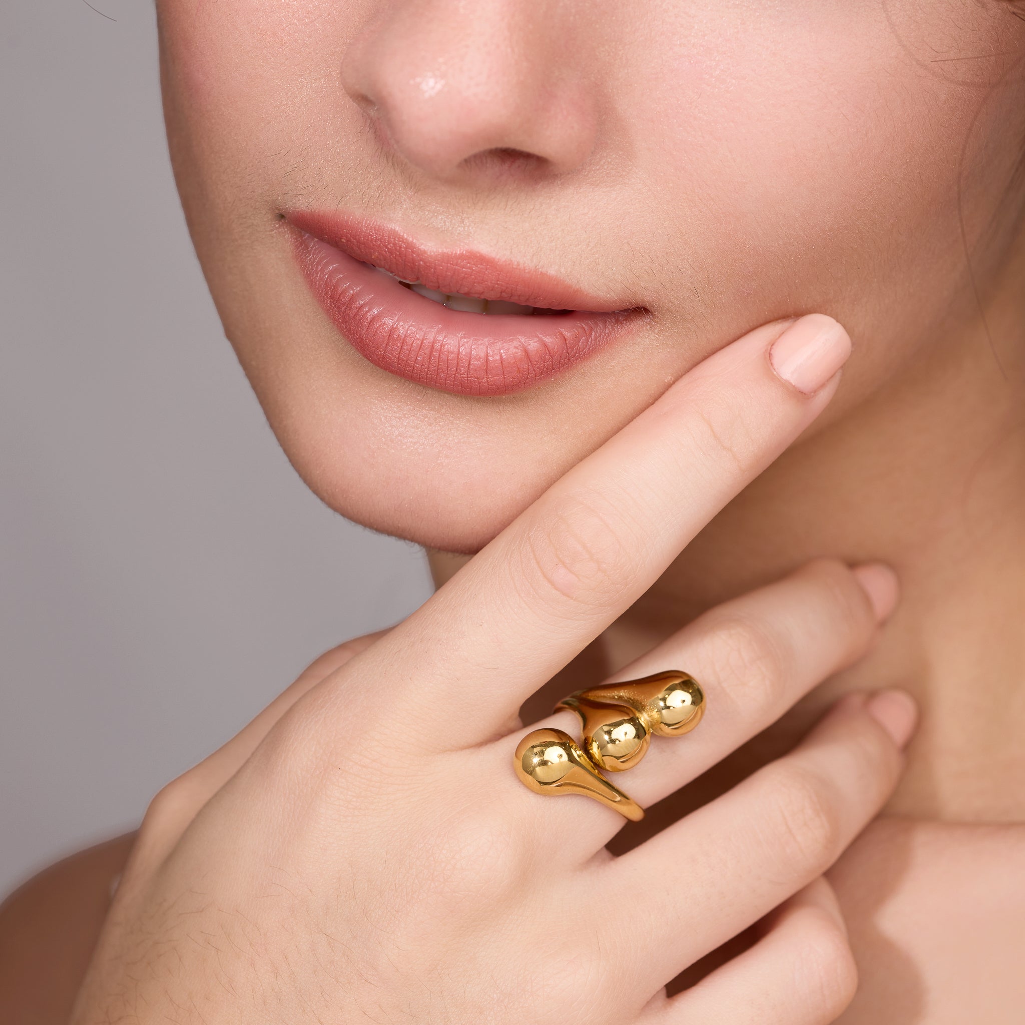 Close-up of a woman's face with a gold ring on her finger against a neutral background