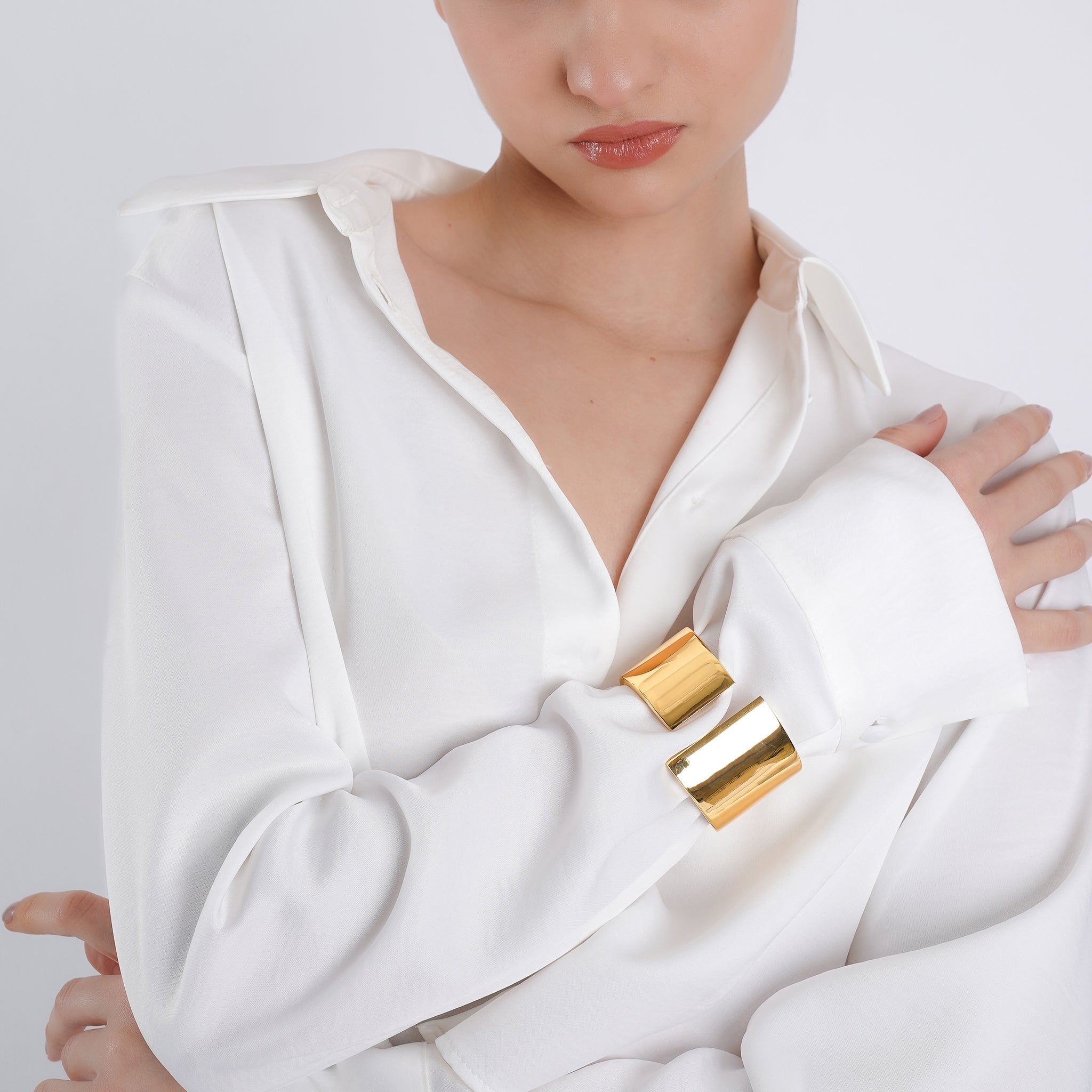 A woman in a crisp white button-down shirt showcasing a bold, polished gold cuff bracelet on her wrist, reflecting light and emphasizing its smooth, structured design.