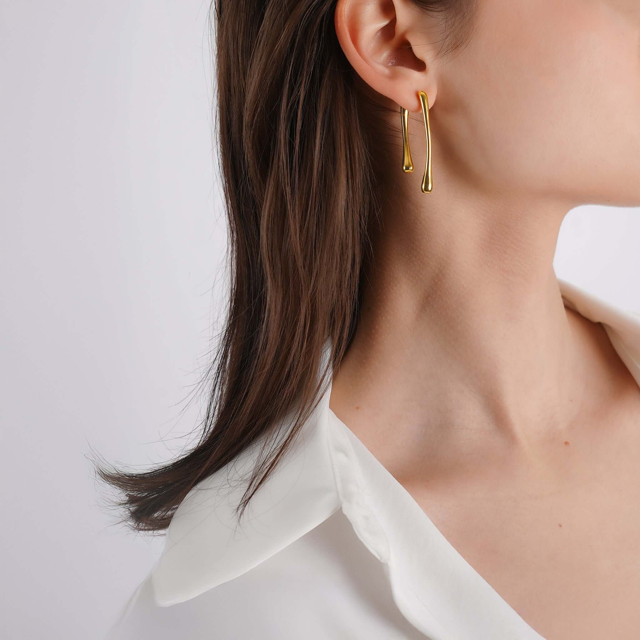 18K gold-plated drop earrings with a fluid, sculptural design elegantly frame the ear. Their organic, asymmetrical shape adds a modern yet timeless touch, perfectly complemented by sleek, straight hair and a crisp white blouse.