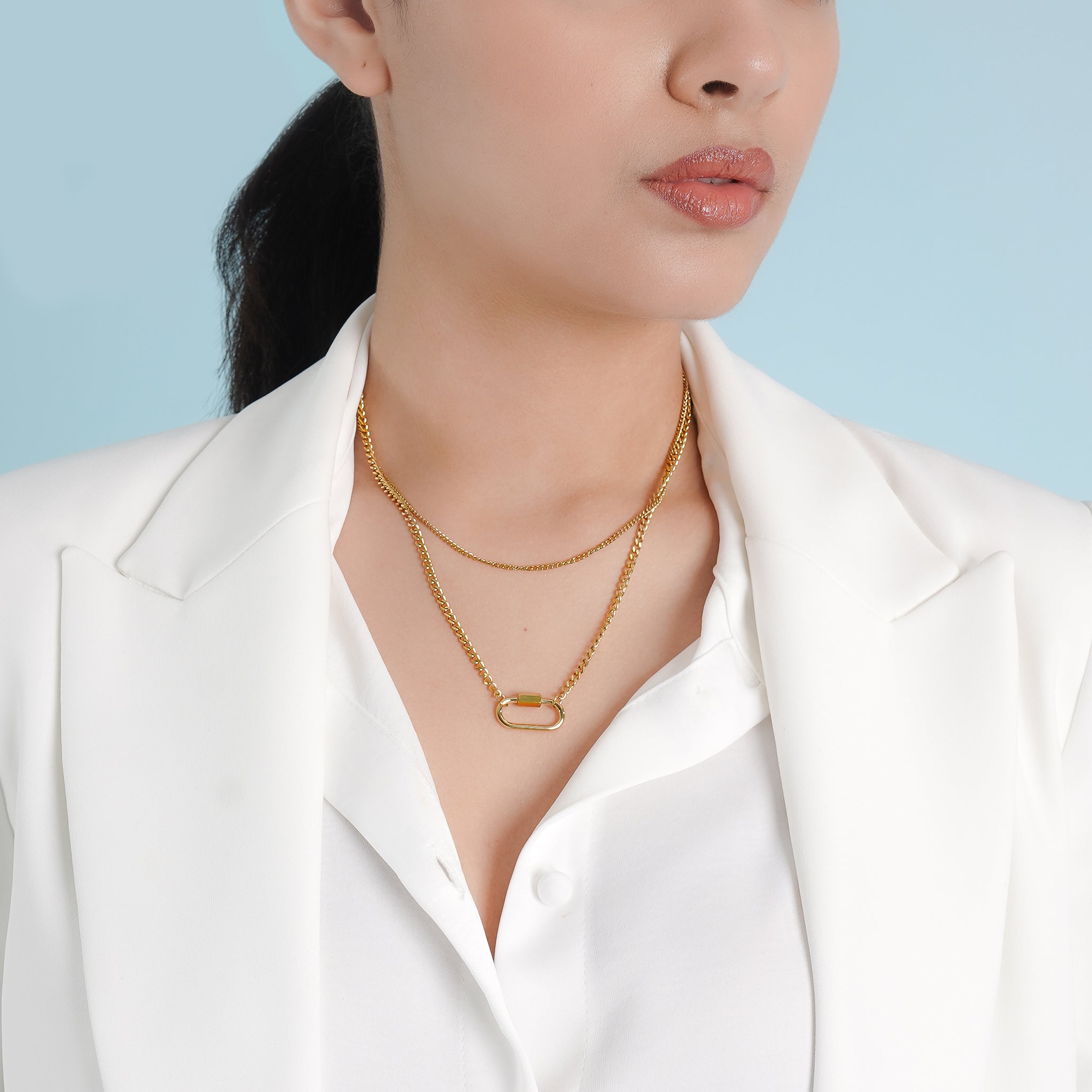 A model wearing the 18K gold-plated layered necklace with interlocking chains and a lock charm, styled elegantly with a white blazer against a soft blue background.