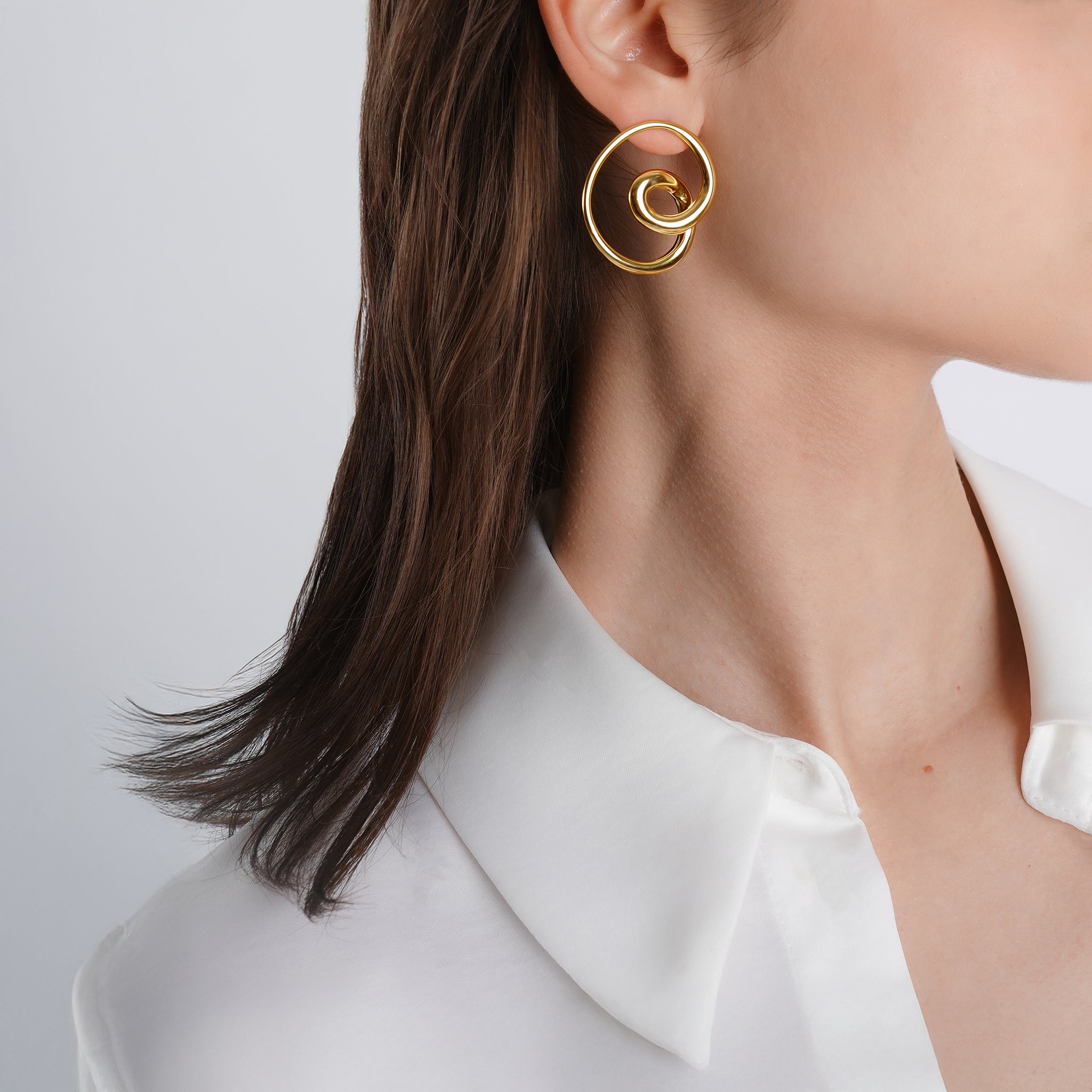 A woman wearing 18k gold-plated circular spiral hoop earrings, styled with a sleek white shirt, highlighting the statement piece against her neutral attire.