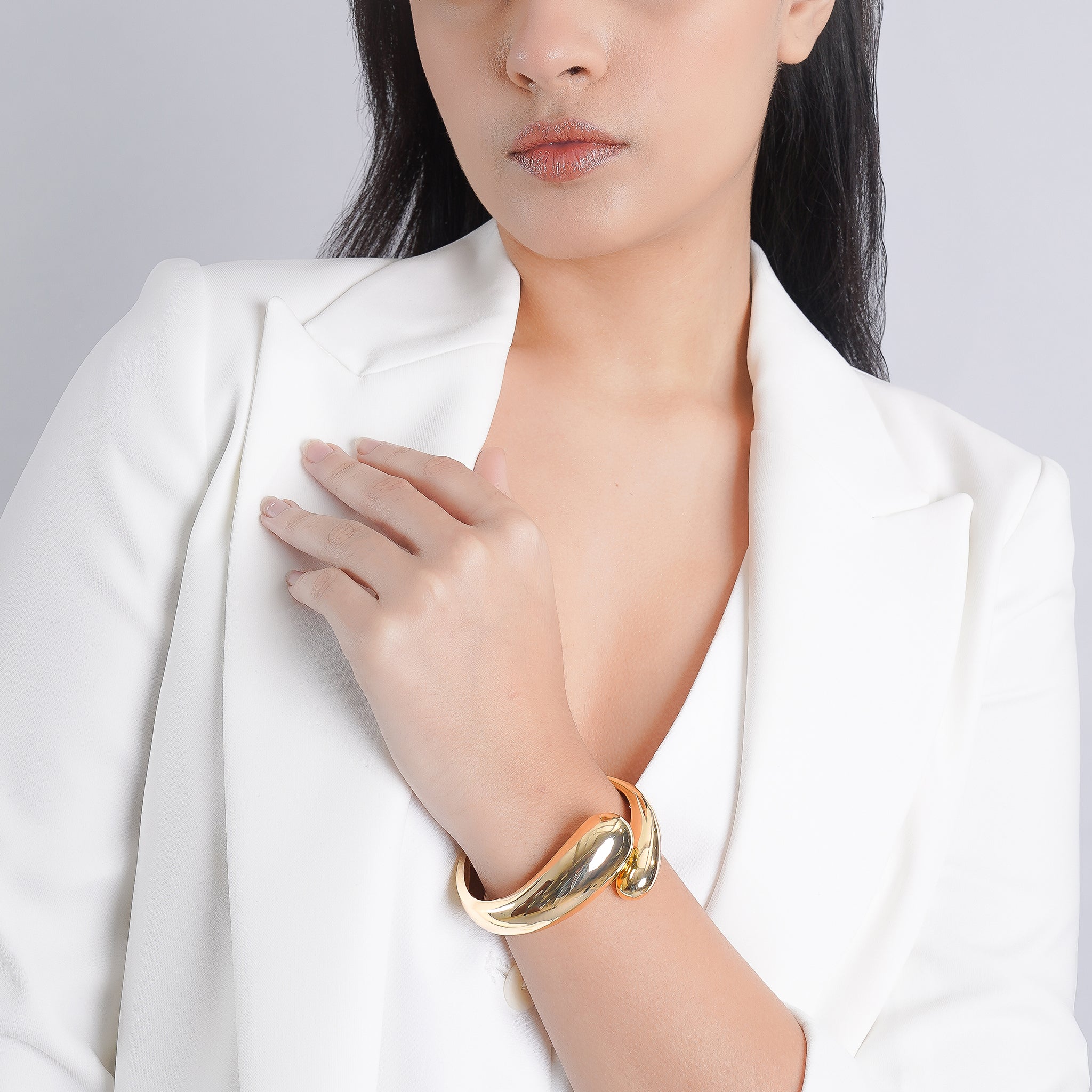  A woman wearing a gold-plated cuff bracelet on her wrist, styled with a white blazer. The reflective, minimalist design highlights its bold yet elegant character, ideal for versatile outfits.
