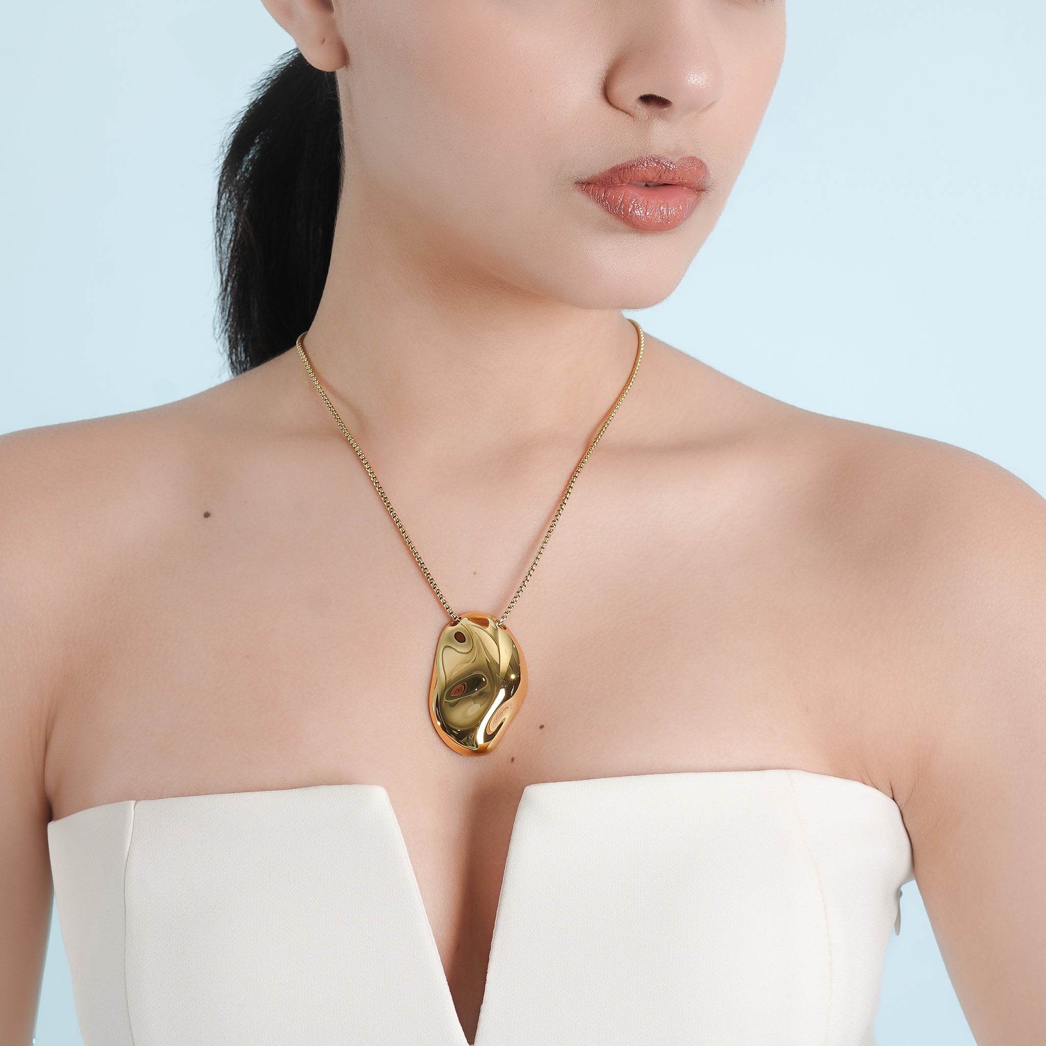 A model wearing the 18K gold-plated necklace with a smooth, sculptural pendant, styled elegantly over a strapless white outfit.