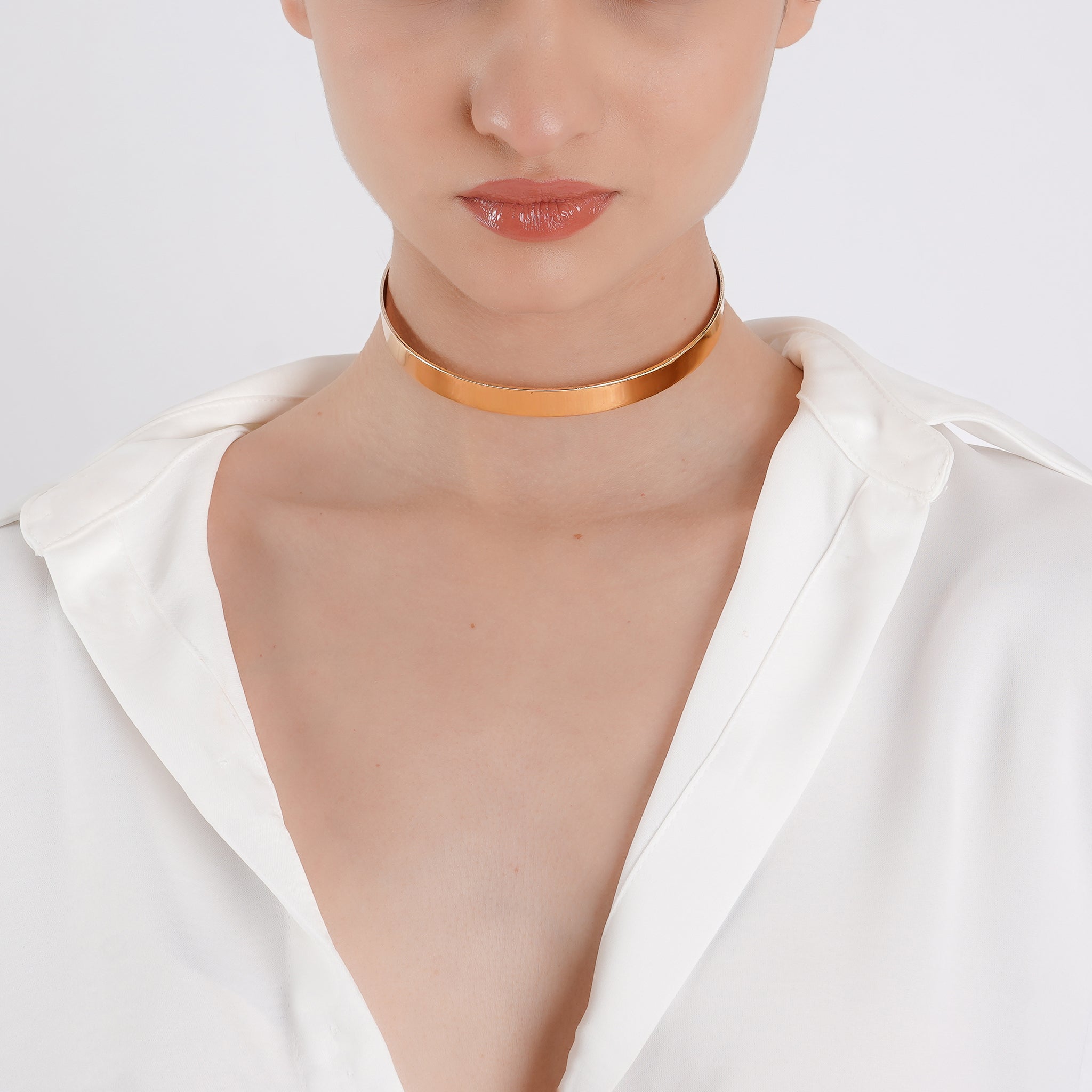 A minimalist, shiny gold choker wraps around the model's neck, accentuating the elegant neckline, paired with a white open-collar blouse for a refined, contemporary look.