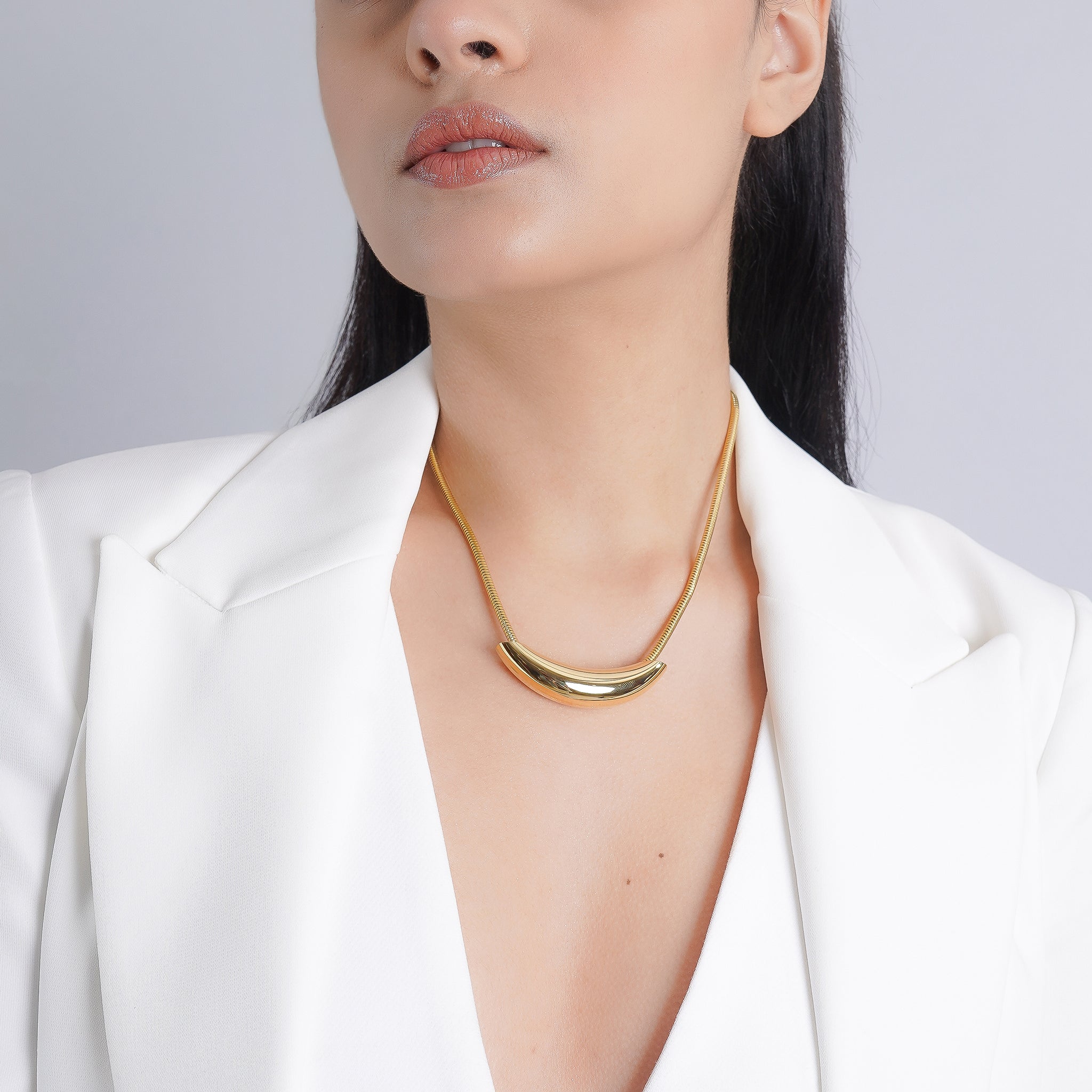 The model elegantly showcases the 18K gold-plated crescent pendant necklace, resting gracefully against the collarbone, complementing a white blazer for a refined and sophisticated look.