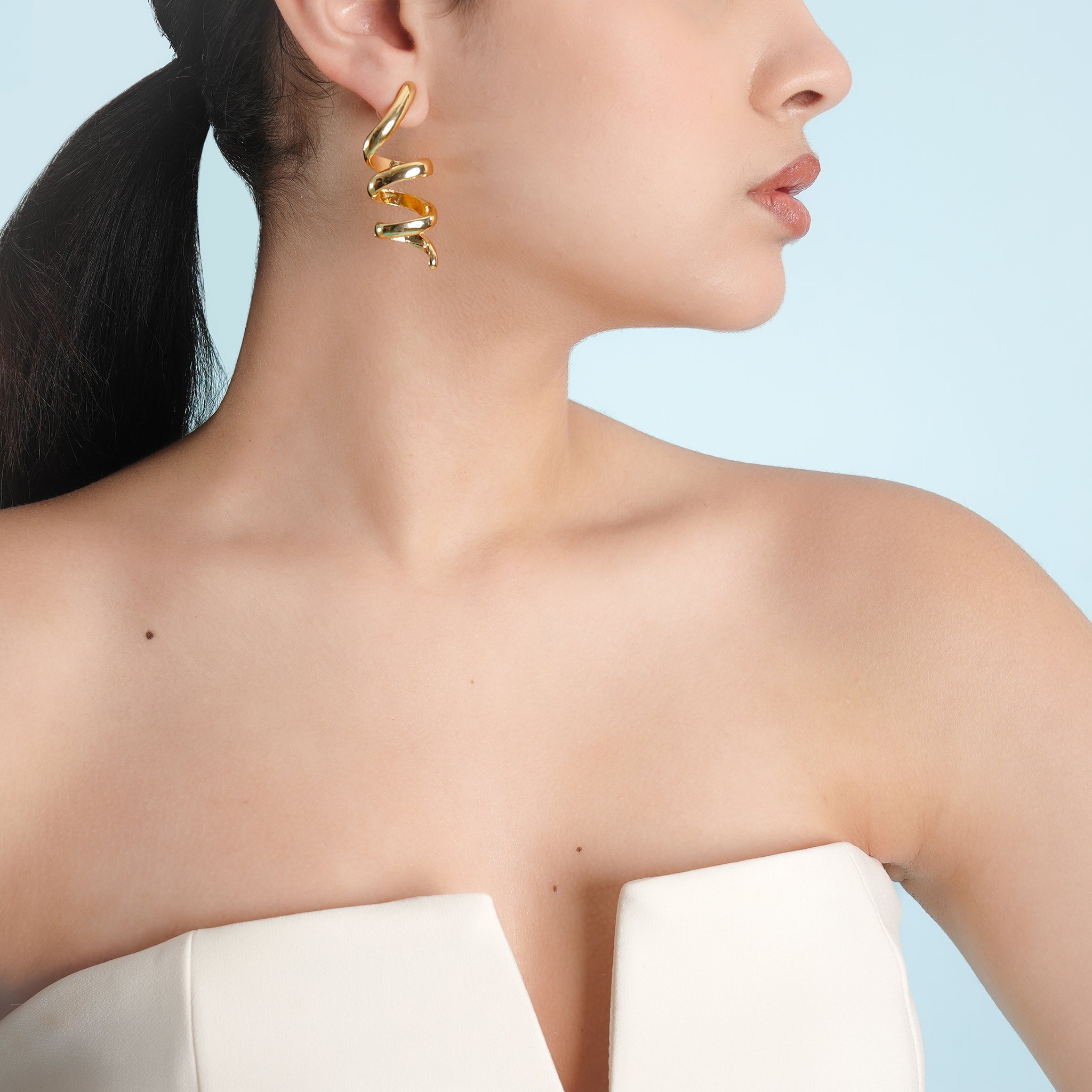 A close-up view of a woman wearing gold-plated spiral earrings, accentuating their dynamic twist design against a minimalist white outfit.
