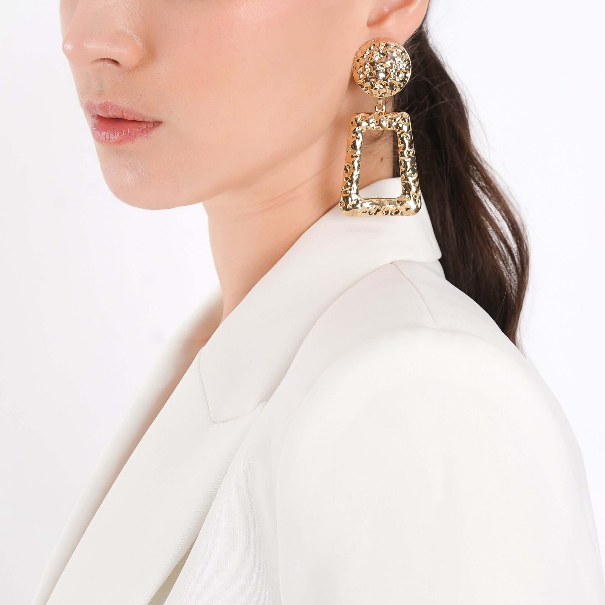  A close-up of a model wearing gold-plated, hammered geometric earrings with circular studs and trapezoid drop pendants, styled elegantly with a white blazer, highlighting the earrings’ statement appeal.