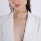 A model wearing the wavy, gold-tone choker against a white blazer, highlighting the necklace’s sleek, bold curves and its modern, sophisticated appeal.