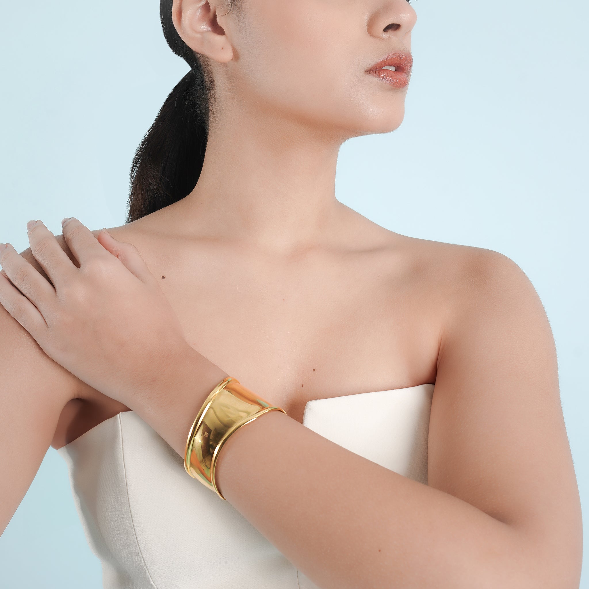 A model wearing the 18K gold polished cuff bracelet on her wrist, paired with a strapless, minimalist white outfit, highlighting the bracelet's bold, reflective design.
