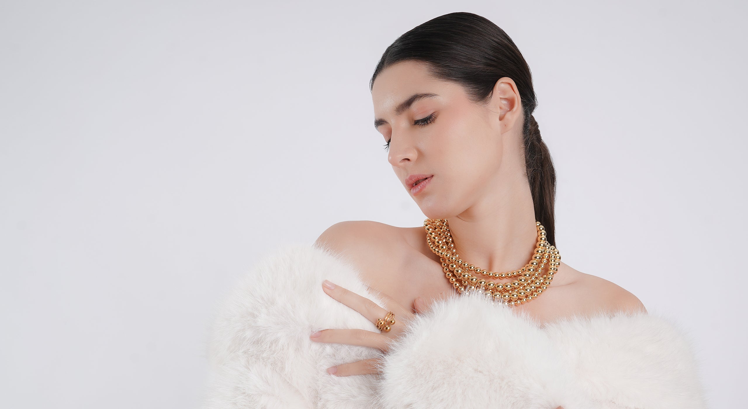 A woman draped in a white faux fur coat poses elegantly, wearing a statement 18K gold-plated beaded necklace and matching ring. Her sleek ponytail and soft makeup enhance the luxurious and refined aesthetic against a clean white background.