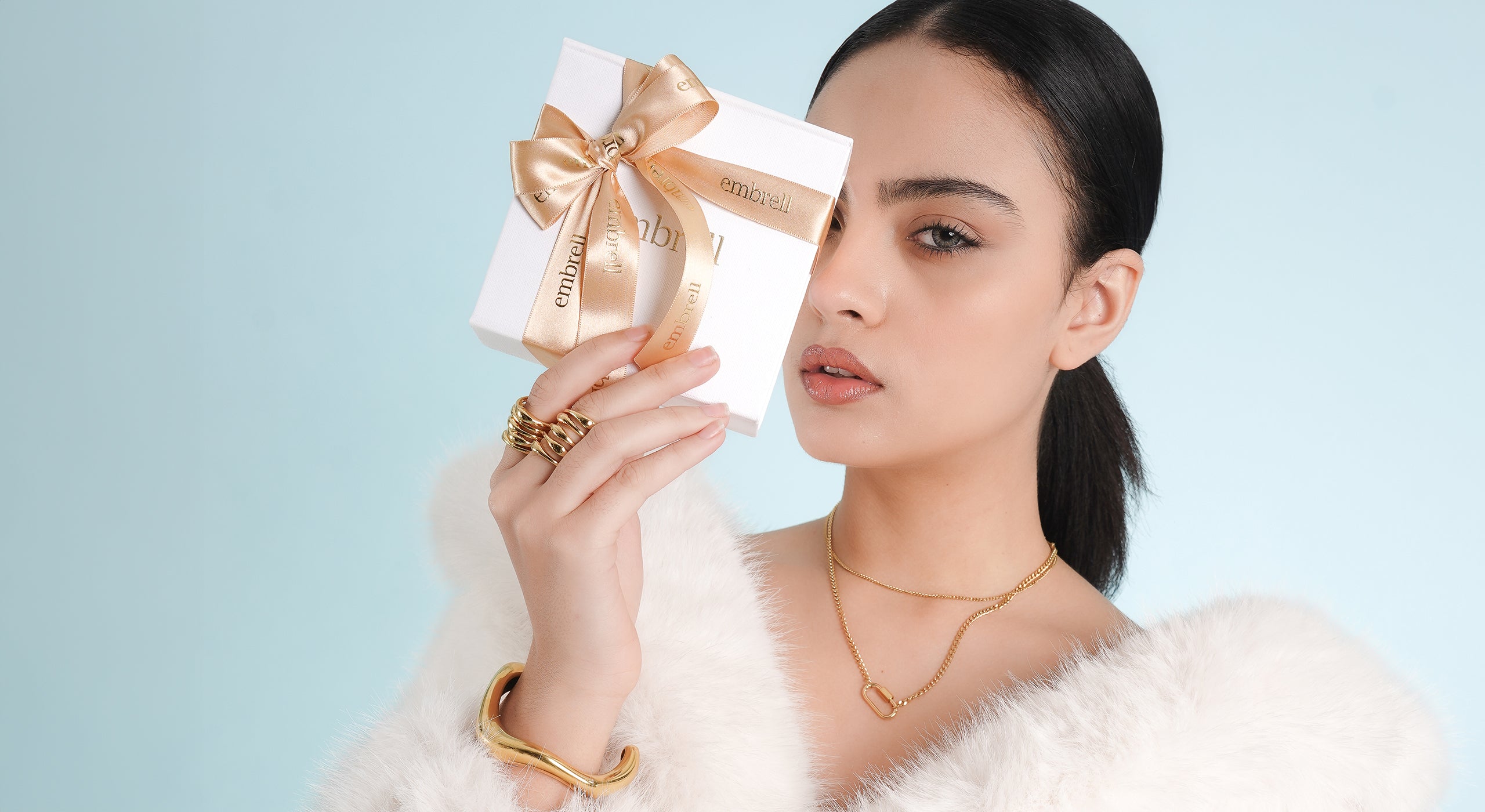 A woman in a white faux fur coat holds an Embrell gift box with a gold ribbon close to her face. She wears layered 18K gold-plated necklaces, statement rings, and a bold cuff bracelet, exuding elegance against a soft pastel background.