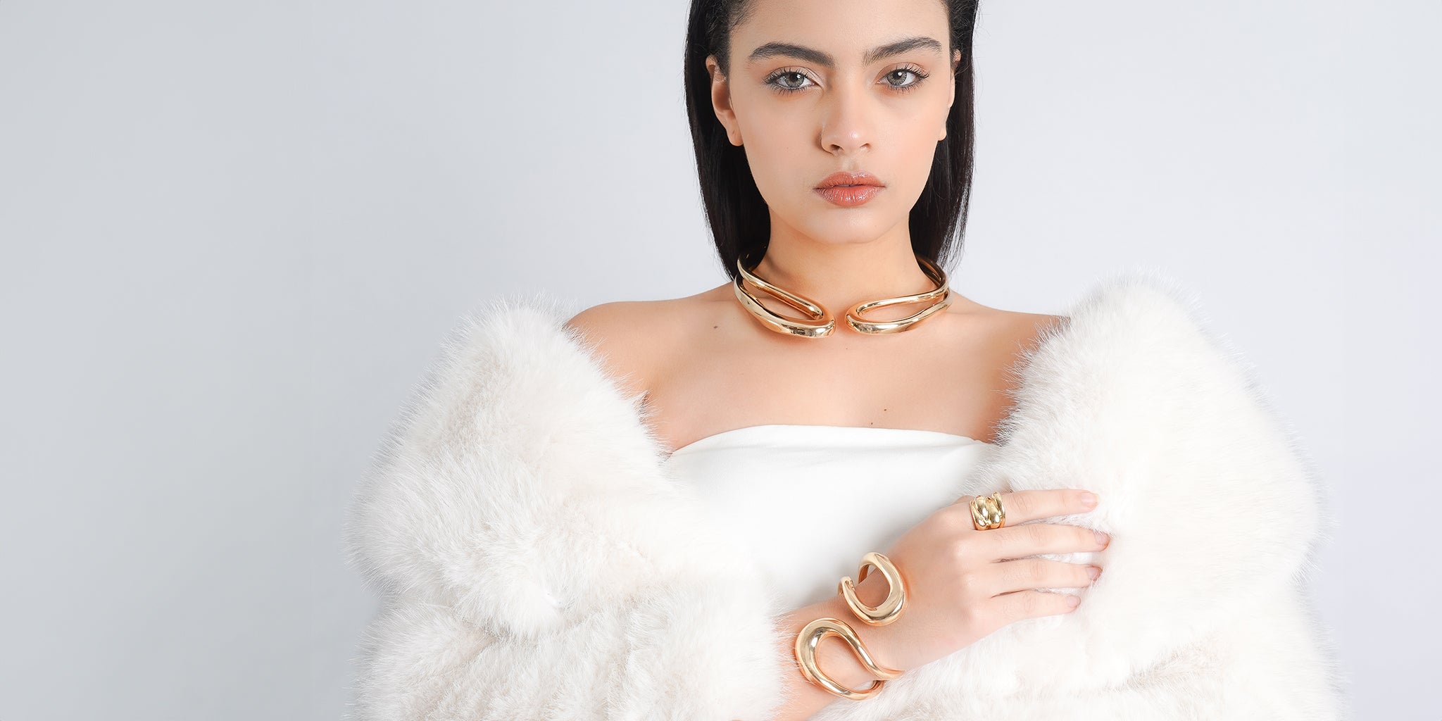 A model elegantly showcasing a luxurious gold jewelry ensemble, featuring a bold statement collar necklace, matching cuff bracelets with fluid curves, and a gold ring with a textured design. The model is dressed in a white fur wrap, highlighting the jewelry’s sophisticated appeal against a soft, minimal background.