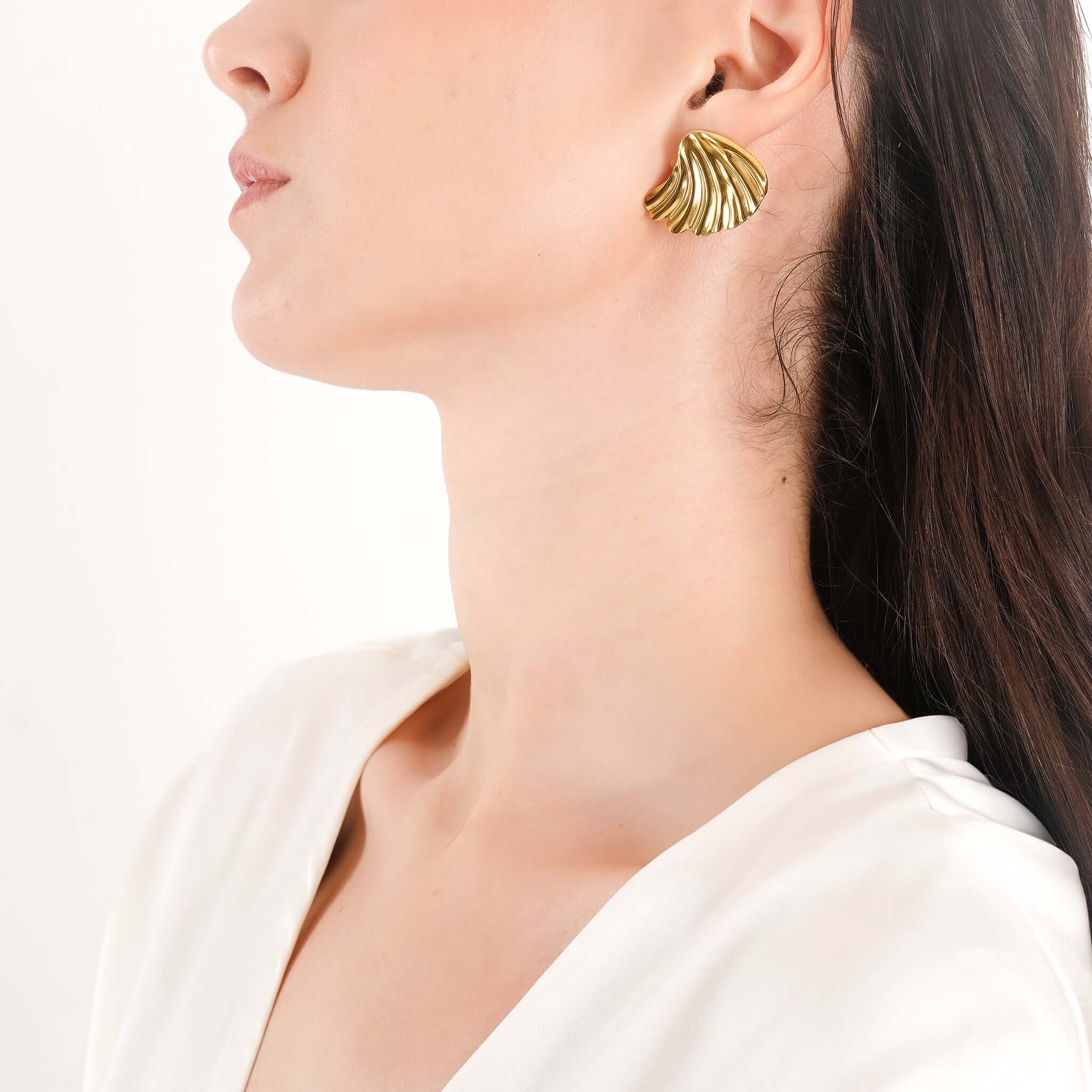 Model wearing 18K gold-plated sculptural shell earrings with a pleated texture, styled with a sleek hairstyle and a white satin top, highlighting the statement piece's bold, elegant charm.