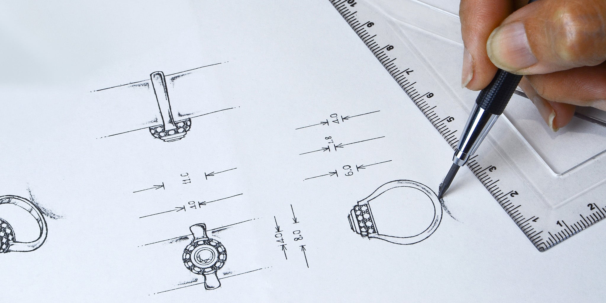 A close-up of a jewelry designer’s hand sketching a detailed technical drawing of a ring, complete with precise measurements. A mechanical pencil and ruler are used to ensure accuracy, highlighting the craftsmanship and precision involved in jewelry design.