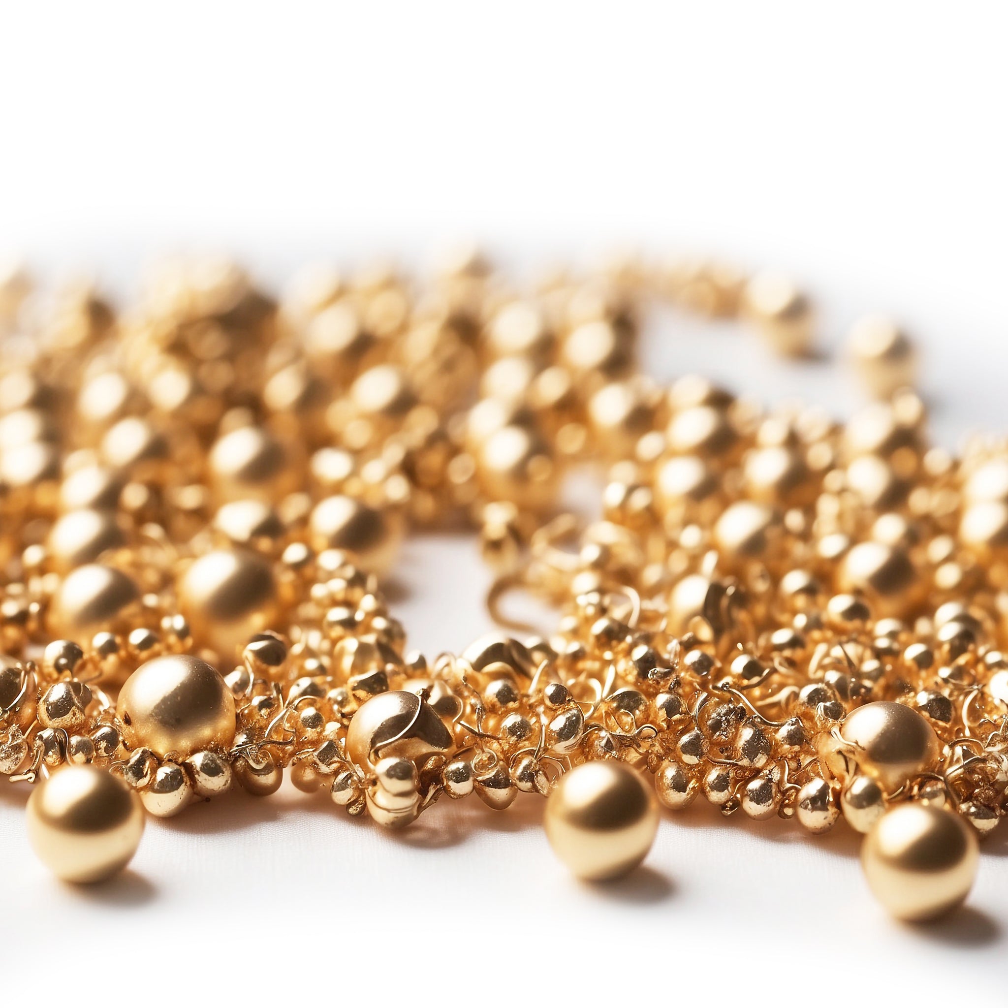 A close-up of an intricate gold bead necklace with various-sized spherical gold beads intertwined in a delicate chain. The lustrous gold finish catches the light, creating a rich, luxurious texture. The blurred background emphasizes the detailed craftsmanship of the jewelry.
