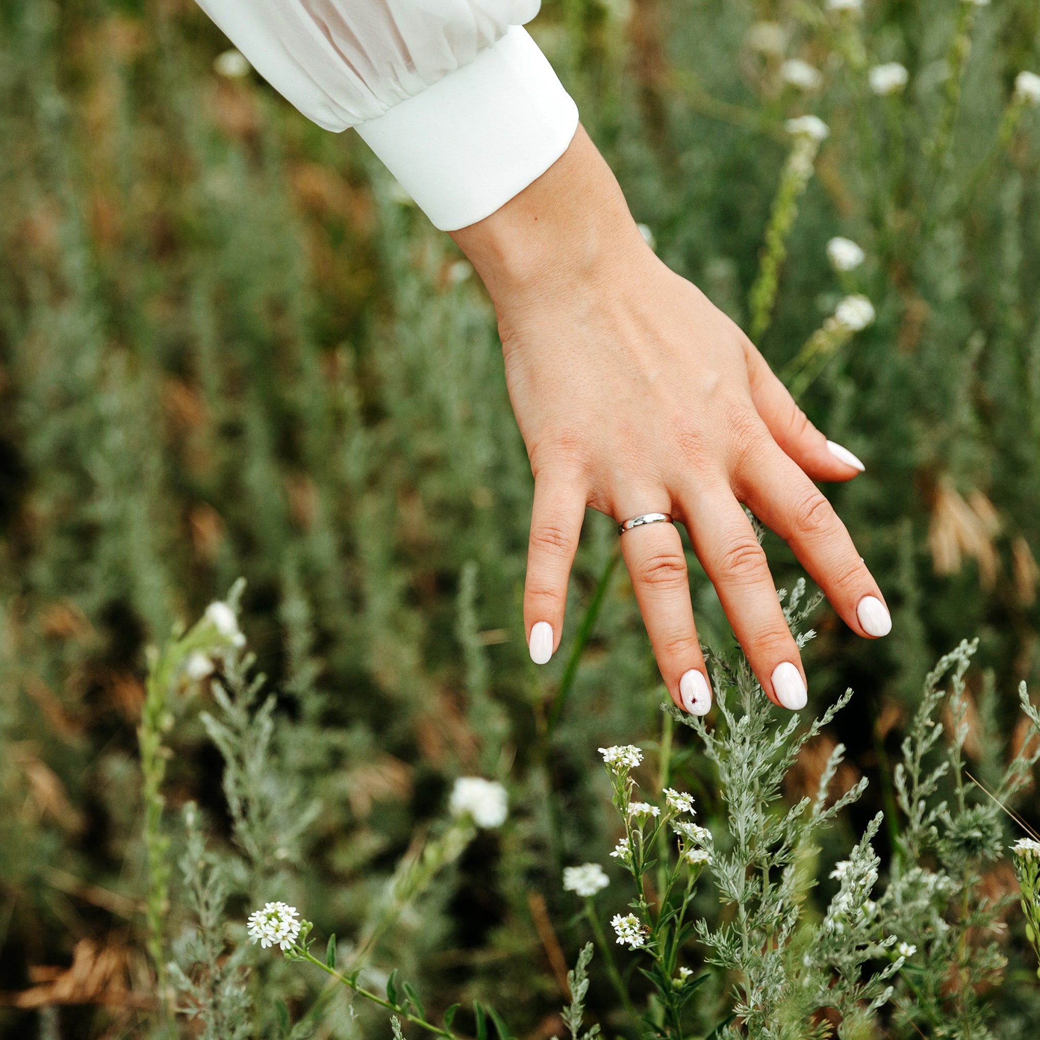 A woman's hand adorned with a sleek silver ring gracefully hovering over delicate white wildflowers. The hand has neatly manicured nails painted in soft white, complementing the elegant white cuff of her blouse. The lush green background adds a natural and serene touch, highlighting the simplicity and beauty of the jewelry.